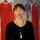 Jaime Black debout devant des robes rouges suspendues à des cintres devant une toile de fond. Cette toile de fond montre l’image d’une forêt de bouleaux dans laquelle d’autres robes rouges sont suspendues.